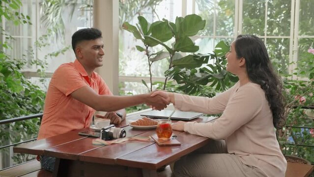 Medium long shot of Indian businessman greeting female colleague with handshake and speaking with her while meeting in cafe with biophilic design interior