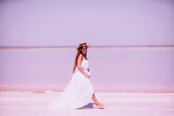 Fototapeta na wymiar Woman in pink salt lake. She walks in a white long dress and hat along the salty white shore of the lake. Wanderlust photo for memory