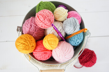 multicolored woolen balls in a light green bowl with crochet hooks on white wooden ground, yellow,...