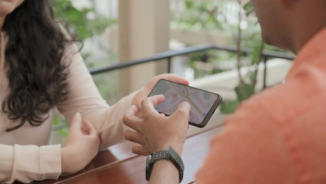 Tilt up over the shoulder shot of man zooming picture or map on smartphone and discussing it with female friend as she holding gadget, both sitting at cafe table