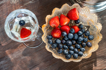 Blueberries and strawberries in a serving plate on a wooden table. Top view. Glass of white wine with berries in out of focus - 590900705