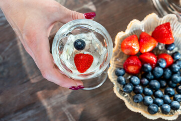 Woman's hand with a glass of white wine and berries. Top view.  Blueberries and strawberries in a serving plate on wooden table in out of focus - 590900712