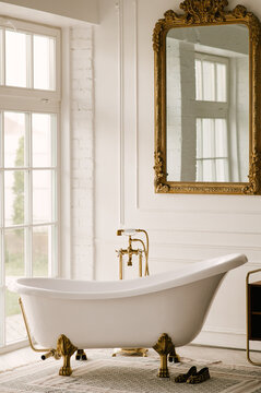 Free-standing bathtub near the window with gold faucets and vintage mirror