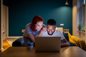 Happy smiling interracial couple man and woman look at laptop screen while spending time together...