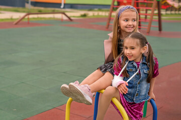 Girl with fractures hand and her friend are sitting on the playground crossbar at the kindergarten.