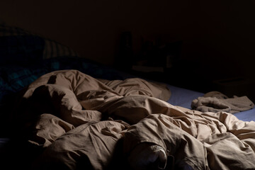 An empty unmade bed in the dark. A crumpled blanket lit by a lamp.