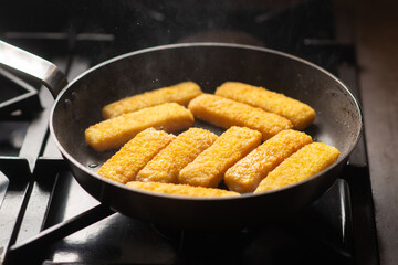 Breaded fish sticks in a frying pan. Preparation of frozen fish sticks. Fast food. Dark background.