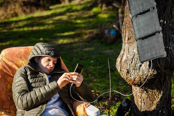 A portable solar battery is hanging from a tree. A man resting in nature with a mobile phone in his hands. Charging phone from solar eco energy.