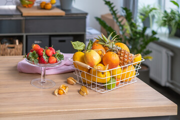 Juicy fruits in a metal basket, strawberries in a glass bowl, physalis and a linen napkin on a light wooden table. A shelf and green houseplants are in the blurred background.