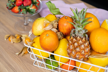 Fototapeta na wymiar Juicy sunny fruits in a white metal basket, fresh red strawberries in a glass bowl, physalis and a linen napkin on a light wooden table.