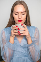 Beautiful young serious woman in light blue dress, with gorgeous long hair, red lips and nails, ring finger, looking at coffee with whipped cream cap.