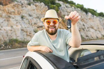 Car driver man smiling showing new car keys and car. Bearded guy driving rented cabrio on summer...