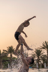Dad and son are playing in the swimming pool. The father tosses the son up.