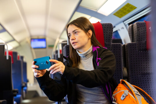 Dark-haired woman enjoying journey in modern comfortable train, looking with interest at picturesque scenery outside window and taking pictures on cellphone