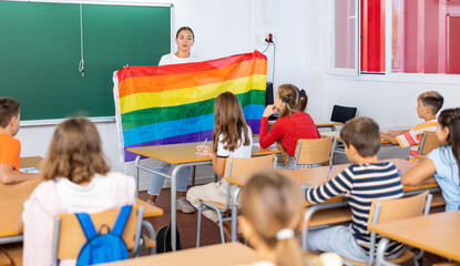 Young woman teacher holding rainbow flag while explaining lesson about LGBTQ people to young girls and boys. - obrazy, fototapety, plakaty