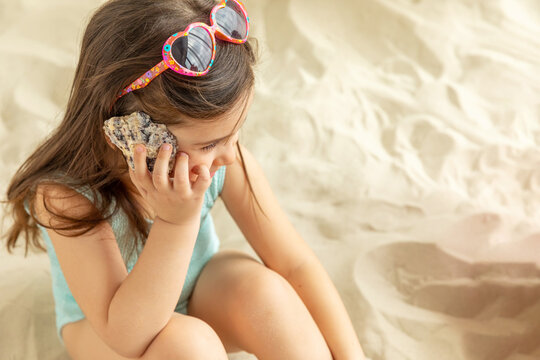 Little girl sits on a sandy beach and holds a large seashell near her ear, space for text