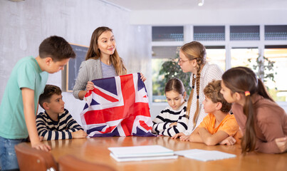Decent teacher showing flag of Great Britain to group of preteen schoolchildren in classroom during...