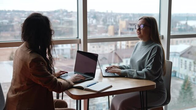 Business ladies discussing ideas, building strategies, cooperating. Women sit in front of each other having laptops on the desk. Panoramic window at backdrop.