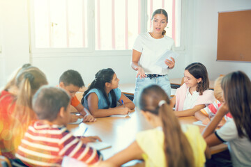 Young female teacher conducting lesson with interested preteen children sitting around common desk in classroom