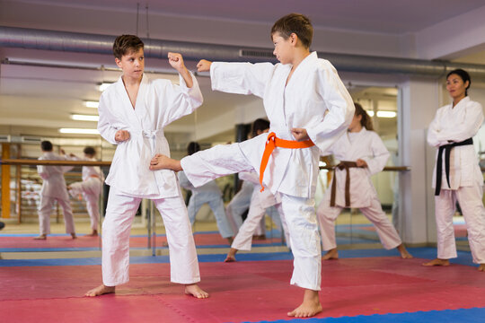 Two boys in kimono sparring together and exercising kicks on group training. Trainer standing beside and watching them.