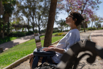 a beautiful girl freelancer in glasses works at a laptop in the summer in the park against the backdrop of palm trees next to a bottle of water