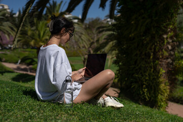 a beautiful girl freelancer in glasses works at a laptop in the summer in the park against the backdrop of palm trees next to a bottle of water