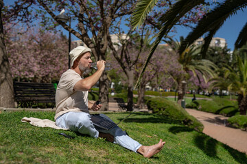 Fototapeta premium bearded male freelancer in the park with a laptop drinks water from a bottle while sitting on the grass with bare feet