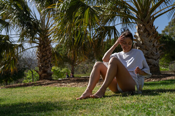 beautiful young girl is resting sitting in the park on the green grass against the backdrop of palm trees on a hot summer day