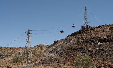 Old, rusty mine carts hanging on a cable between two steel towers in Broken Hill, New South Wales, Australia. One tower stands on a hill; the other below. Deep blue sky.