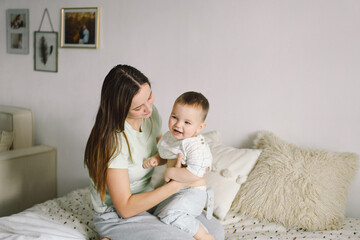 Home portrait of a baby boy with mother on the bed relaxing and playing in the bed at the weekend together, lazy morning, warm and cozy scene. Mom holding and kissing her child. Mother day concept