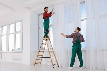 Workers in uniform hanging window curtain and showing thumbs up indoors