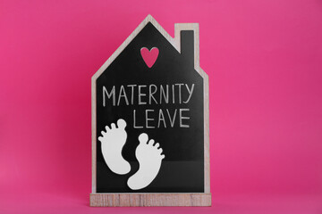 Wooden house figure with words Maternity Leave and paper cutout of baby feet on pink background