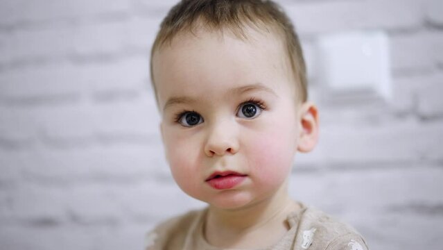 Adorable big-eyed baby boy with mousy hair. Portrait of a calm Caucasian kid focused on something. Close up. Blurred white backdrop.