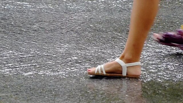 A woman's foot in summer sandals walking slowly on asphalt with water flowing from heavy rain.