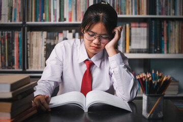Unhappy teenage Asian girl student in school uniform wearing glasses sitting in the library reading...