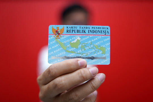 A person showing and holding an Indonesian identity card (KTP) using a red t-shirt and a red background. Selective focus