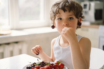 Cute beautiful little boy eating fresh cherry and strawberry. Healthy food, childhood and development. Happy kid at home.