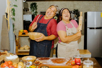 Happy fat asian couple laughing and healthy having fun together kneading and mixing dough to cook homemade pizza in home kitchen. The two of them held wooden utensils and laughed in a good mood.
