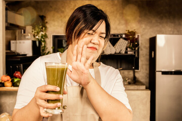 Obese asian woman holding glass of smoothie making her not want to drink organic vegetable juice mixed with fruits that has an unpleasant taste and pungent smell smells in a glass, disgusting face.