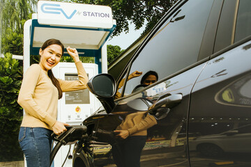 Smiling asian woman raises her hands in delight as the battery of the electric car is nearly full...