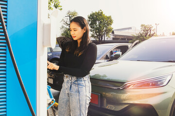 Young asian woman driving an EV standing in front of DC charging station looks at the process of...