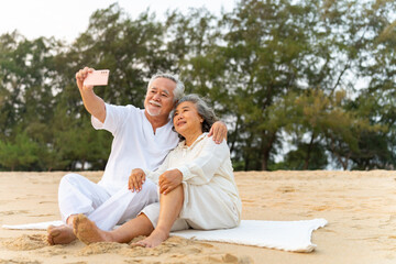 Happy Asian family senior couple using mobile phone taking selfie together during resting on the beach at sunset. Retired elderly people enjoy outdoor lifestyle travel ocean on summer holiday vacation
