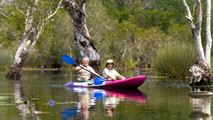 Asian senior couple kayaking together in the lake at mangrove forest on summer vacation. Retired elderly people man and woman have fun outdoor lifestyle travel nature and rowing a boat in the river.