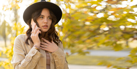 Young woman talking on cell phone in autumn park with shocked expression. Banner.