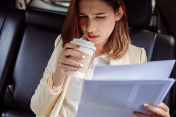 Businesswoman sitting in the back seat of the car with a paperwork in her hand and drinking coffee.