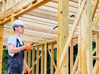 Laborer constructing wooden frame house near forest. Man treating woods, applying fire retardant using sprayer, while dressed in protective suit, helmet. Concept of modern eco-friendly construction.