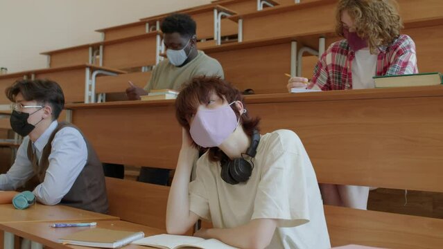 Group of multi-ethnic college or university students wearing protective masks sitting at desks in lecture hall listening to professor and making notes