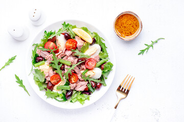 Nicoise salad with tuna, red tomatoes, boiled eggs, green beans and olives on plate, white table background, top view