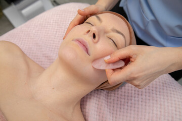 Obraz na płótnie Canvas Closeup woman lying on couch, receive Chinese gua sha quartz lifting facial massage. Cosmetologist hands antiage massage