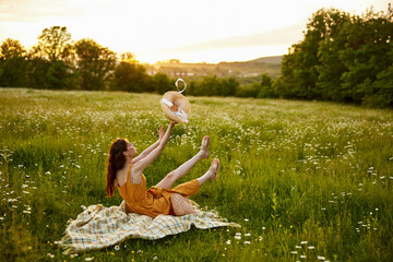 a happy woman in an orange dress sits on a plaid in a chamomile field at sunset and throws her straw hat into the air